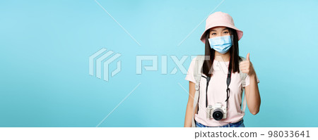 Cheerful asian woman in medical face mask, holding camera, showing thumbs up, travelling abroad, going on vacation. Tourist standing over blue background 98033641