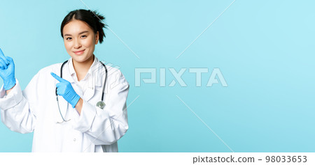 Smiling female doctor, physican in medical uniform and sterile gloves, pointing fingers left at promo, clinic logo, standing over blue background Smiling female doctor, physican in medical uniform and sterile gloves, pointing fingers left at promo, clinic logo, standing over blue background 98033653