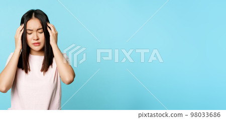Asian young girl with headache, massaging her head with troubled face expression, painful migraine, standing over blue background 98033686