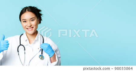 Smiling asian woman therapist, physician with stethoscope shows thumbs up, standing over blue background Smiling asian woman therapist, physician with stethoscope shows thumbs up, standing over blue background 98033864