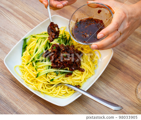 Close to the teaspoon and the hands of a person putting mushroom and minced meat sauce to the noodles with fresh cucumbers Close to the teaspoon and the hands of a person putting mushroom and minced meat sauce to the noodles with fresh cucumbers 98033905