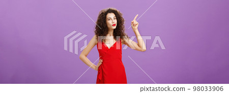 Woman ready to shine on dance floor in elegant red dress with sensual makeup and curly hairstyle pointing at upper left corner smirking with delight and self-assured expression over purple wall Woman ready to shine on dance floor in elegant red dress with sensual makeup and curly hairstyle pointing at upper left corner smirking with delight and self-assured expression over purple wall 98033906