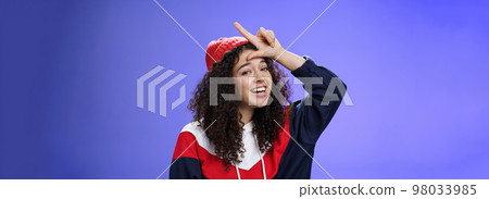 Girl triumphing and mocking friend as winning showing loser gesture on forehead and laughing having fun standing pleased and happy over blue background, smiling at camera joyfully Girl triumphing and mocking friend as winning showing loser gesture on forehead and laughing having fun standing pleased and happy over blue background, smiling at camera joyfully 98033985