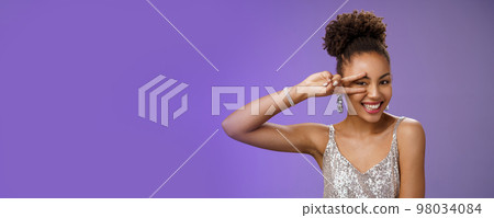 Slim stylish beautiful young elegant african american woman in trendy silver dress brilliant accessorise smiling broadly show peace victory gesture near eye feel lucky amused, blue background 98034084