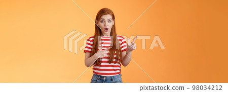 Wow awesome. Impressed excited beautiful redhead woman look astonished explain amazing news, stare camera thrilled talking about advertisement, pointing left upbeat impressed, orange background 98034212