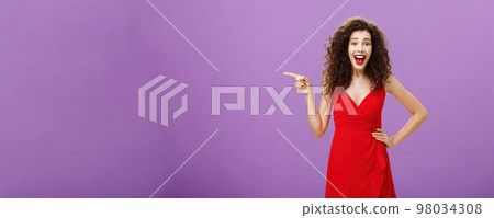 Portrait of entertained and amused joyful european woman with curly hairstyle in red evening dress laughing from amusement and joy pointing left being entertained posing over purple background 98034308