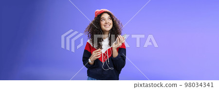 Dreamy charming and happy kind girl recalling nice memories as listening nostalgic song in earphones holding smartphone looking at upper left corner with broad smile over blue background Dreamy charming and happy kind girl recalling nice memories as listening nostalgic song in earphones holding smartphone looking at upper left corner with broad smile over blue background 98034341