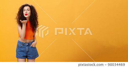 Stylish flirty and confident female with curly hairstyle and red lipstick smiling sensually bending towards camera sending gentle loving kiss at camera with palm near mouth posing near orange wall Stylish flirty and confident female with curly hairstyle and red lipstick smiling sensually bending towards camera sending gentle loving kiss at camera with palm near mouth posing near orange wall 98034469