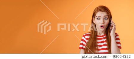 Amused thrilled redhead girl found out incredible news, talking phone, hold smartphone ear, stare camera surprised amazed, telling about awesome promo, stand astonished orange background 98034532