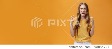 Portrait of irritated cute european female squeezing hands in fists with anger frowning and making hateful face standing annoyed and pissed over orange background with furious look 98034727