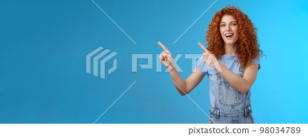 Hey check this out. Enthusiastic redhead curly stylish summer girl inviting beach party pointing upper left corner directing awesome location smiling broadly discuss awesome promo blue background 98034789
