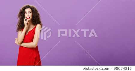 Silly and timid charming woman with dark curly hair in stylish evening dress with red lipstick biting finger looking cute at upper right corner thoughtful imaging or dreaming about desirable thing Silly and timid charming woman with dark curly hair in stylish evening dress with red lipstick biting finger looking cute at upper right corner thoughtful imaging or dreaming about desirable thing 98034815