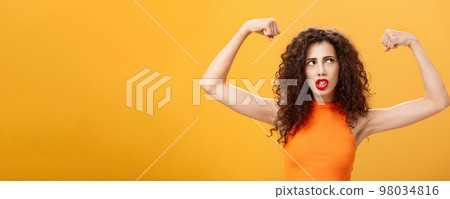 Woman feeling powerful and strong raising hands with clenched fists making intense face being working out in gym showing muscles and biceps looking at upper right corner posing over orange background 98034816