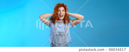 Excited amused joyful redhead curly girl having amazing perfect news touch hair astonished stare camera thrilled cheerful overjoy smiling broadly toothy carefree grin denim overalls blue background 98034817