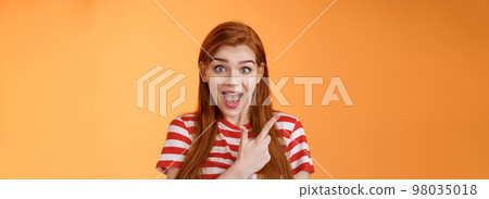 Excited female redhead hear impressive sale offer pointing upper left corner, drop jaw astonished stare camera full disbelief surprise, discuss interesting promotion, stand orange background 98035018
