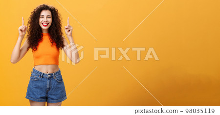 Indoor shot of friendly and cute happy relaxed female with red lipstick and curly hairstyle wearing cool cropped top and denim shorts smiling broadly pointing up posing over orange background Indoor shot of friendly and cute happy relaxed female with red lipstick and curly hairstyle wearing cool cropped top and denim shorts smiling broadly pointing up posing over orange background 98035119