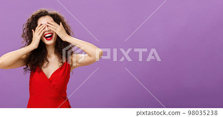 Wife waiting for husband present gift for anniversary closing eyes with palms and smiling broadly peeking through fingers being charmed and excited standing in seductive red dress over purple wall Wife waiting for husband present gift for anniversary closing eyes with palms and smiling broadly peeking through fingers being charmed and excited standing in seductive red dress over purple wall 98035238