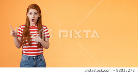 Wow awesome. Impressed excited beautiful redhead woman look astonished explain amazing news, stare camera thrilled talking about advertisement, pointing left upbeat impressed, orange background 98035278