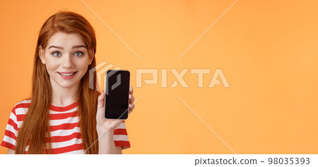 Close-up lovely caucasian redhead girl showing smartphone screen orange background, smiling cute, recommend application, promote app, online offer sale, introduce mobile phone 98035393