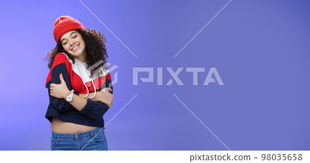 Sweet tender and sensual curly-haired woman in red stylish beanie hugging herself with closed eyes and nostalgic smile leaning on shoulder feeling warmth and tenderness as recalling loving memories Sweet tender and sensual curly-haired woman in red stylish beanie hugging herself with closed eyes and nostalgic smile leaning on shoulder feeling warmth and tenderness as recalling loving memories 98035658