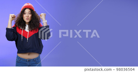 Charming young 20s girl with curly hair in warm beanie raising hands as belly showing under sweatshirt triumphing, celebrating awesome win folding lips from joy and excitement over blue background 98036504
