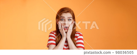 Close-up shocked speechless redhead woman, drop jaw gasping, touch cheeks embarrassed shook, look pity shame, stare camera astonished, surprised hear disturbing news, orange background 98036505