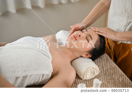 Relaxed Asian woman lying on massage table with eyes closed, receiving facial massage 98038713