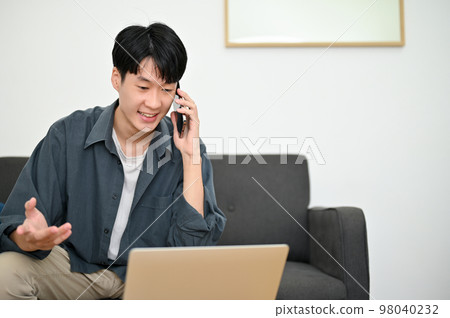 Happy young Asian man looking at laptop screen, talking on the phone with his colleagues 98040232