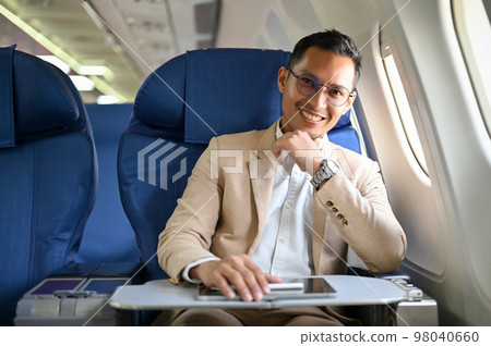 Handsome Asian businessman sits at the window seat in business class, hand on chin, smiling Handsome Asian businessman sits at the window seat in business class, hand on chin, smiling 98040660
