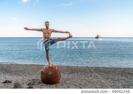 Man doing yoga exercise outdoors standing on old rusty floating marine mine on the beach with rocky shore and sea background. Healthy lifestyle, pollution, nature protection, war and peace concept. Man doing yoga exercise outdoors standing on old rusty floating marine mine on the beach with rocky shore and sea background. Healthy lifestyle, pollution, nature protection, war and peace concept. 98042248