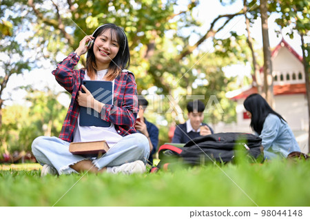Charming Asian female university student wearing headphones, holding tablet, sitting on grass 98044148