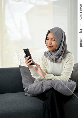 Charming young Asian Muslim woman wearing hijab using her smartphone while relaxing on sofa Charming young Asian Muslim woman wearing hijab using her smartphone while relaxing on sofa 98044696