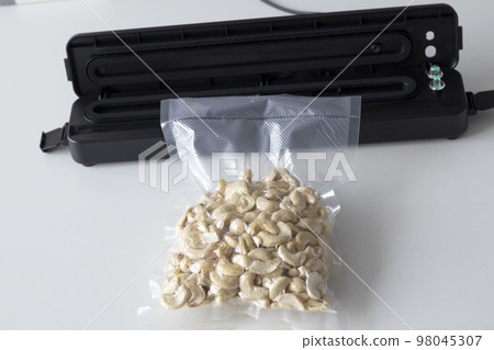 Vacuum packaging machine and vacuum bagged nuts. Device for packing food on a white table with a plastic cashew bag. Vacuum packaging machine and vacuum bagged nuts. Device for packing food on a white table with a plastic cashew bag. 98045307