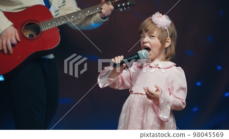Little girl on stage in vintage dress, she sings into microphone and dances, her father plays acoustic guitar. Color music is shining and smoke is billowing. Father and Child Day, performance on stage Little girl on stage in vintage dress, she sings into microphone and dances, her father plays acoustic guitar. Color music is shining and smoke is billowing. Father and Child Day, performance on stage 98045569