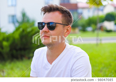 Young male man portrait. European caucasian white young student in sunglasses on green grass lawn on town street, park. Lifestyle Headshot portrait, tourist, businessman Young male man portrait. European caucasian white young student in sunglasses on green grass lawn on town street, park. Lifestyle Headshot portrait, tourist, businessman 98047085