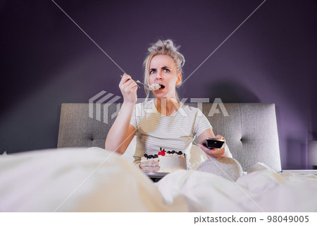 Emotionally stressed, shocked woman eating cake and holding a remote control while watching TV in bed. Scary horror movie or terrible news, scare in her eyes Overeating, Jamming of negative emotions 98049005