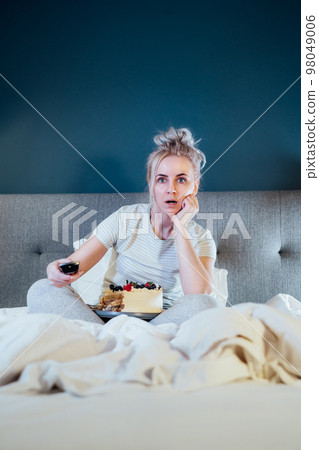 Emotionally stressed, shocked woman eating cake and holding a remote control while watching TV in bed. Scary horror movie or terrible news, scare in her eyes Overeating, Jamming of negative emotions. 98049006