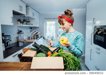 Online home food delivery. Woman checking her online order list on her phone. Cardboard box with fresh vegetables and fruits standing on the kitchen table. Local farmer food. Start of a healthy life. Online home food delivery. Woman checking her online order list on her phone. Cardboard box with fresh vegetables and fruits standing on the kitchen table. Local farmer food. Start of a healthy life. 98049032