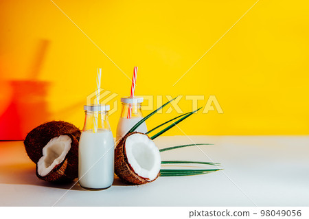 Bottles with coconut non-dairy milk drink and fresh coconuts on bright yellow background with color light. Trendy coctails. Healthy vegan plant base coconut milk. Selective focus. Copy space 98049056