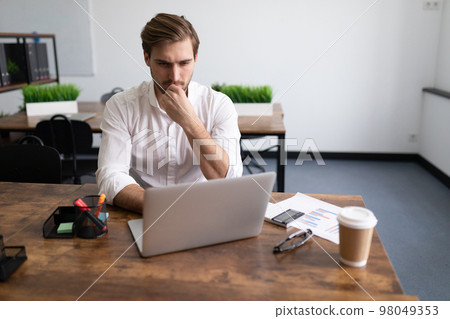 serious businessman in the office behind a laptop solves a complex business problem 98049353