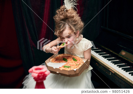 Portrait of cute little girl, child in image of medieval royal person sitting at the piano and emotionally eating pizza 98051426