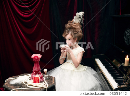Portrait of cute little girl, child in image of medieval royal person sitting at the piano and drinking milkshake 98051442