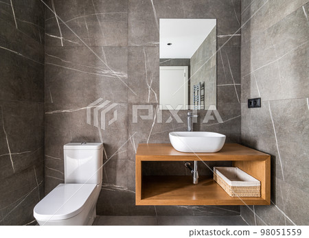 Dressing room with walls made of dark gray marble with white touches, small ceramic oval sink on wooden cabinet and mirror on wall that reflects radiator and front door to room. Stylish bathroom. Dressing room with walls made of dark gray marble with white touches, small ceramic oval sink on wooden cabinet and mirror on wall that reflects radiator and front door to room. Stylish bathroom. 98051559