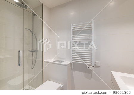 Bathroom in light colors with glossy ceramic tile walls and bright lighting. The bathroom has a shower with a glass railing and radiator for drying towels. Bathroom in light colors with glossy ceramic tile walls and bright lighting. The bathroom has a shower with a glass railing and radiator for drying towels. 98051563