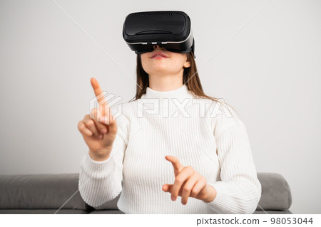 Happy woman in VR glasses moves hand sitting on comfortable couch in apartment living room. Young housewife enjoys modern technology at home Happy woman in VR glasses moves hand sitting on comfortable couch in apartment living room. Young housewife enjoys modern technology at home 98053044
