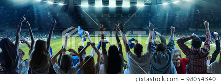 Back view of football, soccer fans emotionally cheering their team at crowded stadium at evening time. Diverse group. Concept of sport, hobby, leisure time, football 98054340