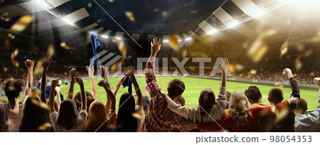 Back view of football, soccer fans cheering their team, holding flag at crowded stadium at evening time. Concept of sport, hobby, leisure time, football 98054353