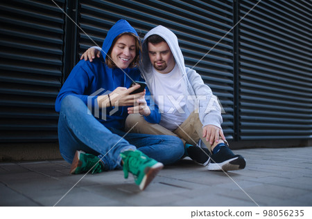 Man with down syndrome resting outdoor with his friend, scrolling smartphone. Concept of friendship and integration people with disability into society. Man with down syndrome resting outdoor with his friend, scrolling smartphone. Concept of friendship and integration people with disability into society. 98056235
