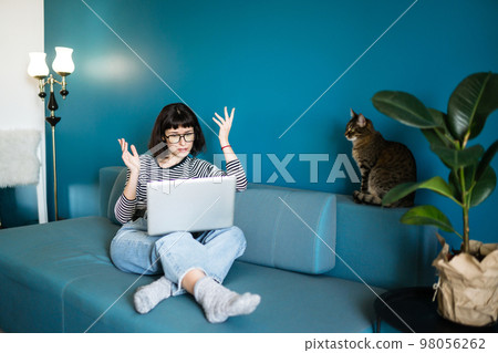 Young woman working with laptop and with a cat sitting in modern living room at home. 98056262