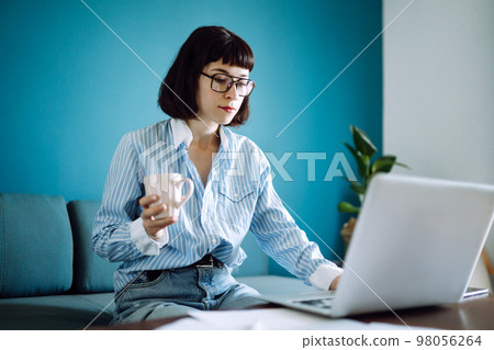 Young woman freelancer sit on modern sofa and work on laptop. Education online. Working from home. 98056264